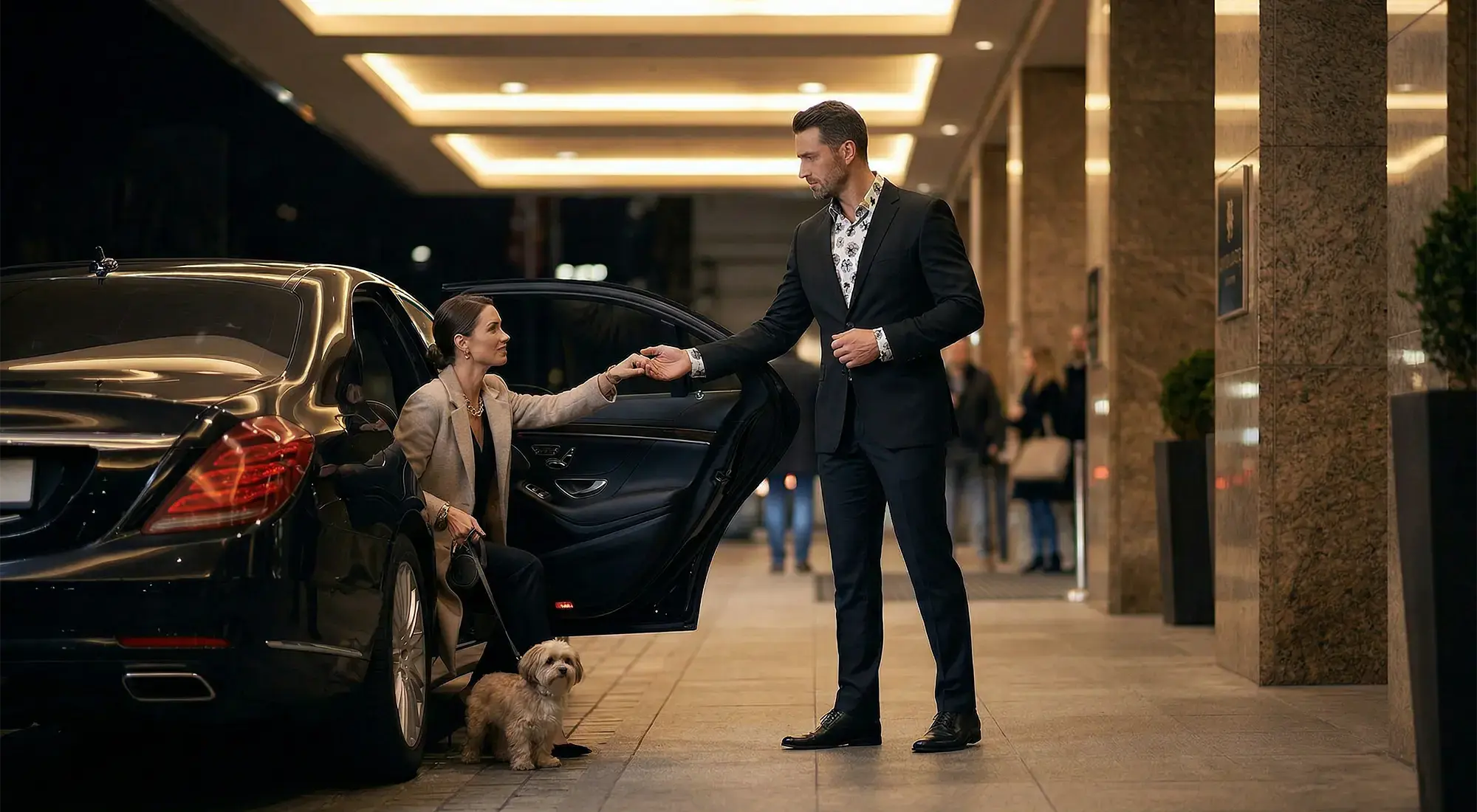 A chauffeur helps a lady with a dog to get out of the car. The chauffeur is wearing a black suit and underneath it the GERMENS OSTSEEBUHNE shirt.