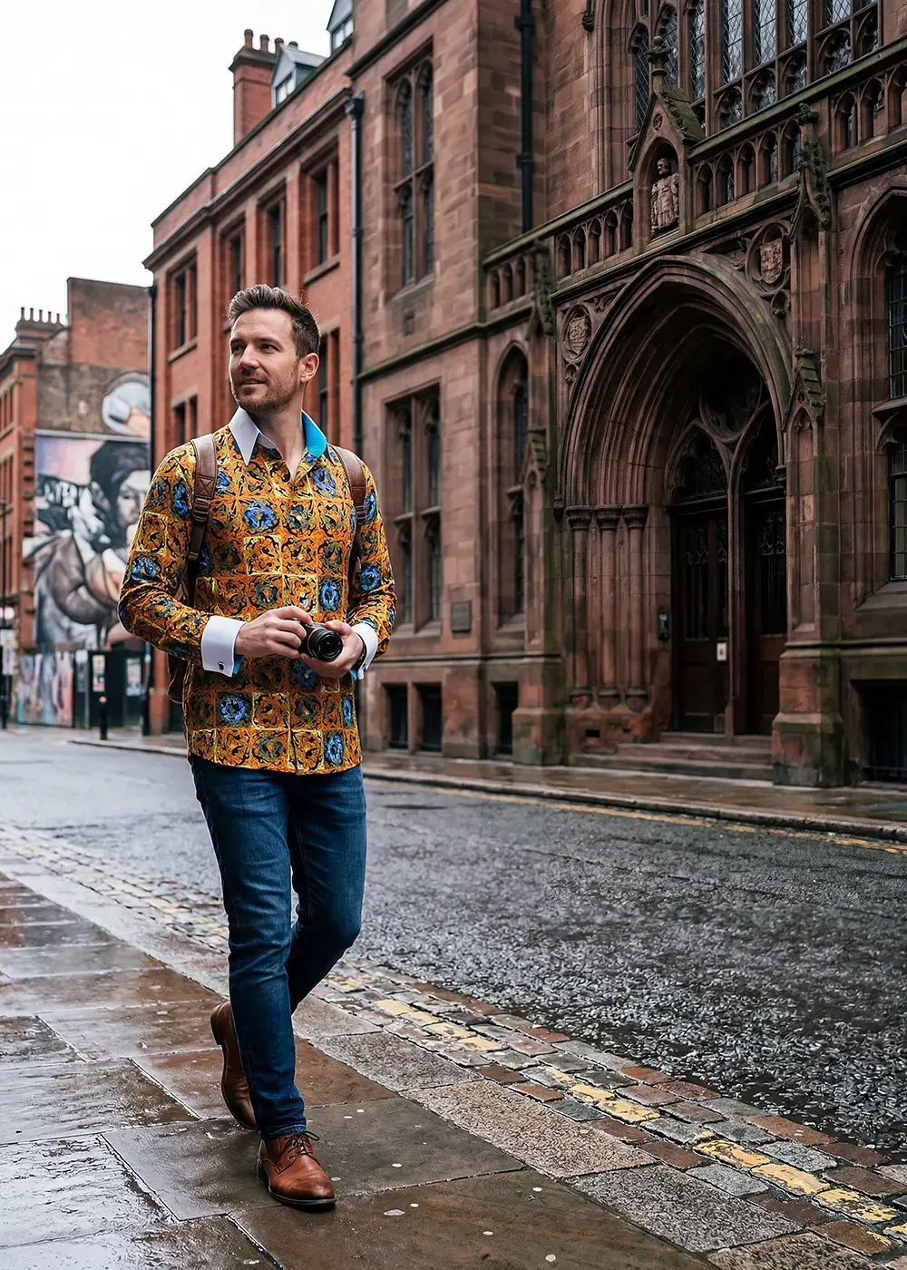 A man walks through Manchester with a camera. He is wearing the orange CUCA shirt from GERMENS.
