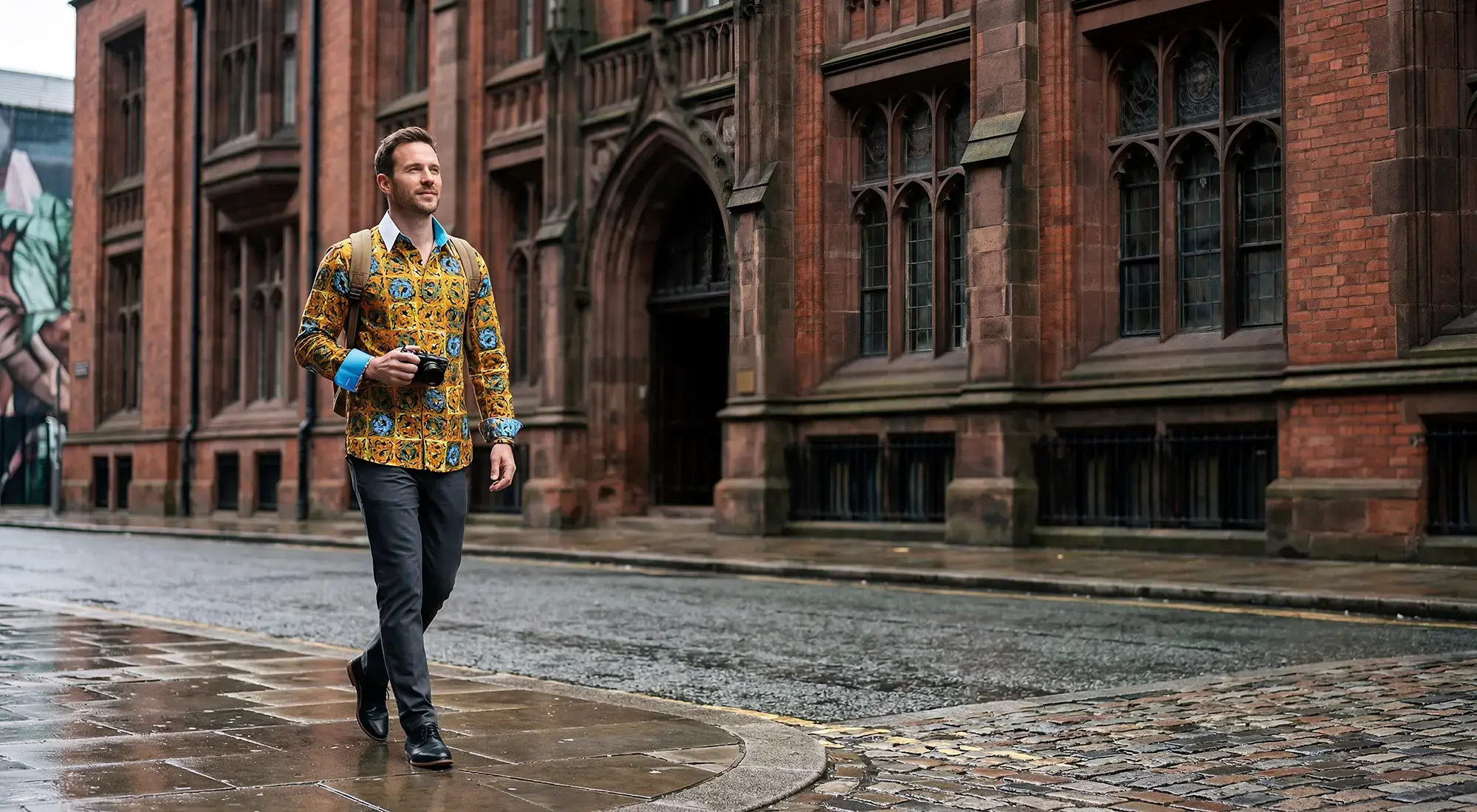 A man walks through Manchester with a camera. He is wearing the orange CUCA shirt from GERMENS.