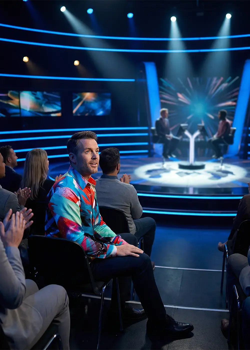 A man is sitting in the audience of a TV show. He is wearing the eye-catching GERMENS shirt THE TIME TRAVELING.